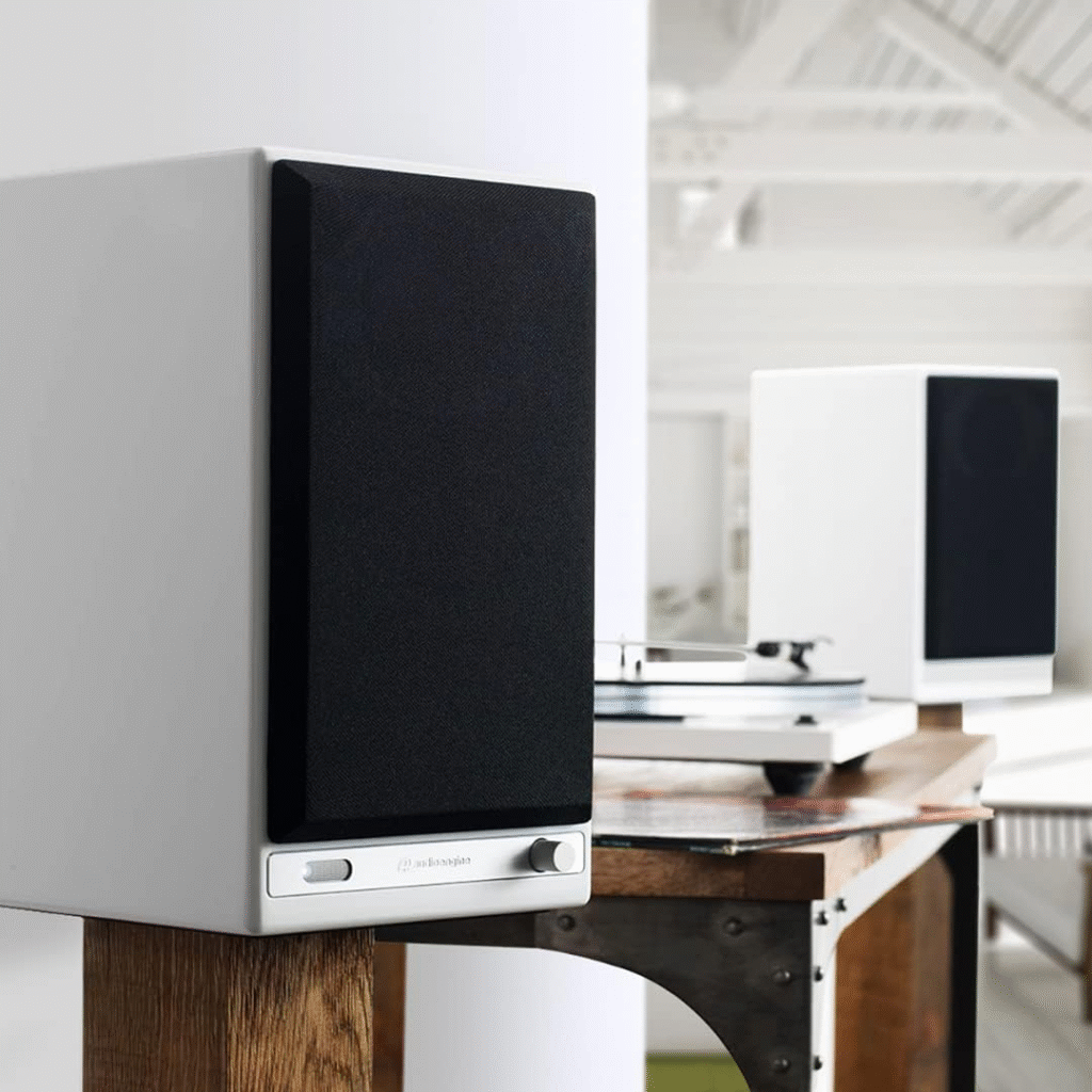 Modern white bookshelf speakers with black front grilles placed on a wooden table in a bright, minimalist room, with a turntable partially visible in the background
