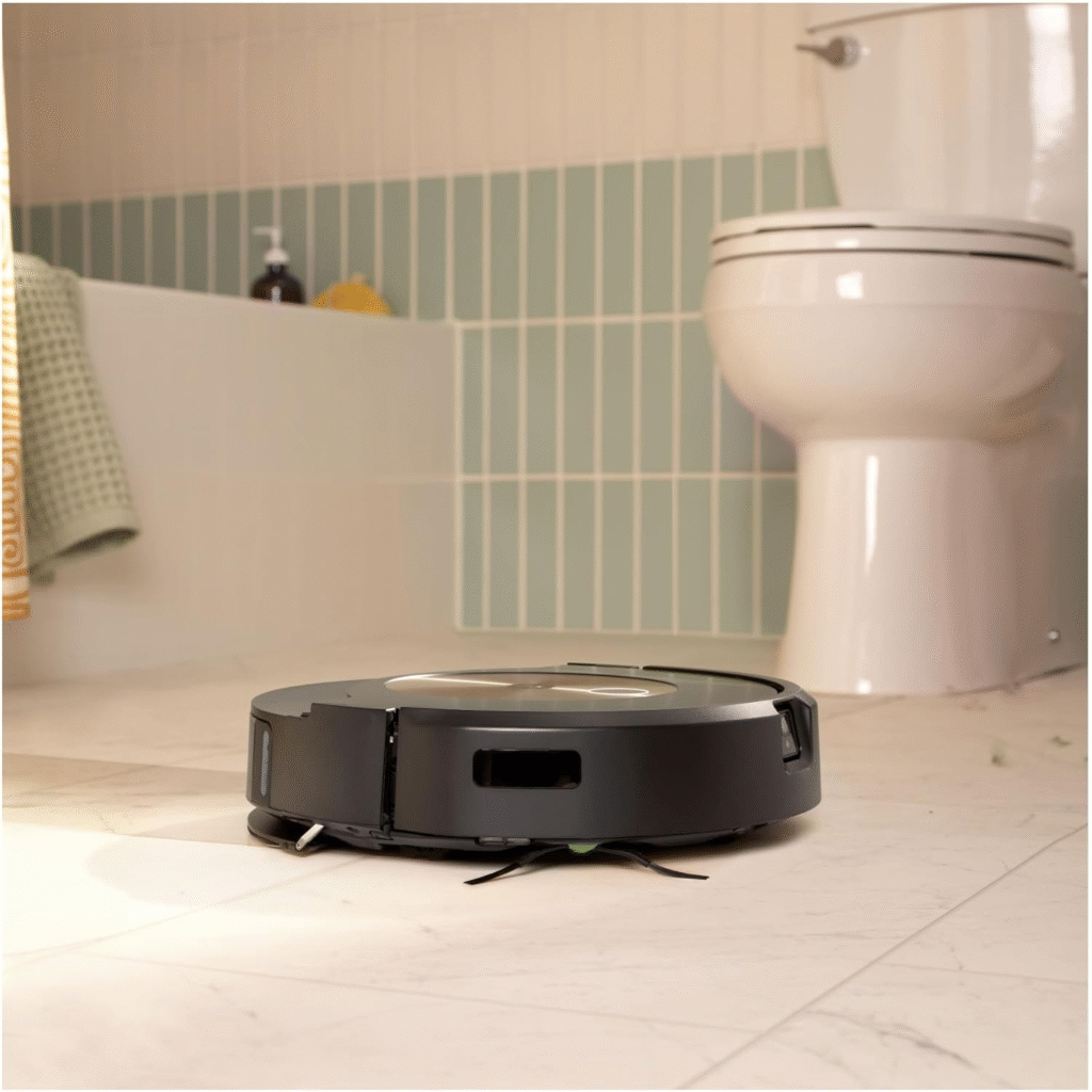 iRobot Roomba vacuum cleans light tiles in a bathroom, with a toilet and tub in the blurred background.